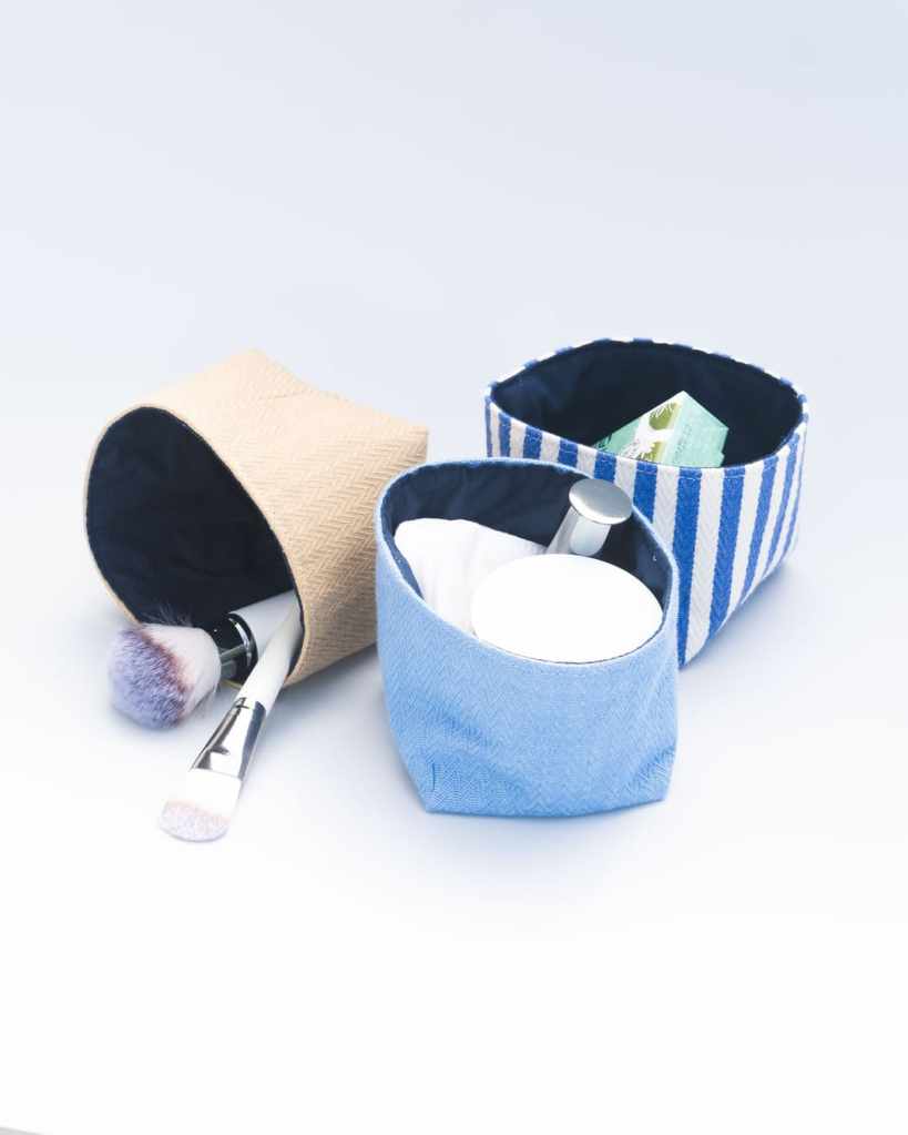 Trois pochons en toile basque, un beige, un bleu unis et un bleu à rayures, disposés sur un fond clair, contenant des produits de beauté.